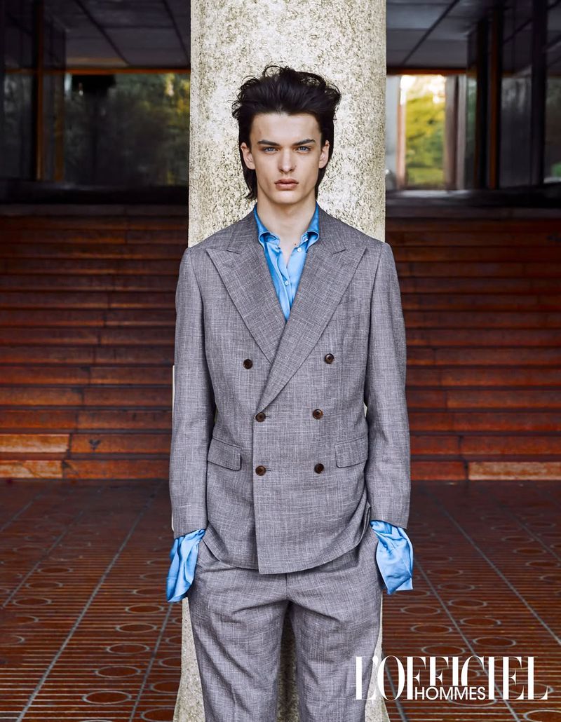 Model Tristan Watkins in a grey double-breasted suit and blue shirt standing before a stone pillar for L'Officiel Hommes Ukraine
