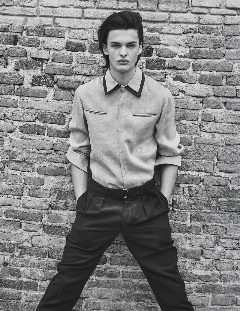 Model Tristan Watkins leaning against an old brick wall, wearing a collared button-down shirt and dark trousers.