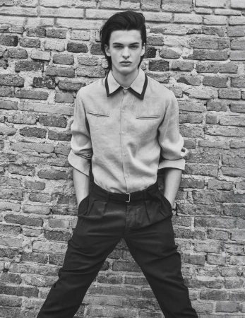 Model Tristan Watkins leaning against an old brick wall, wearing a collared button-down shirt and dark trousers.
