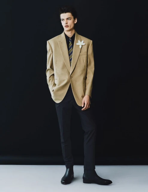 Elegant man in beige blazer, black shirt, and tie strikes a pose against a dark backdrop. Fashion editorial shot with a focus on style.