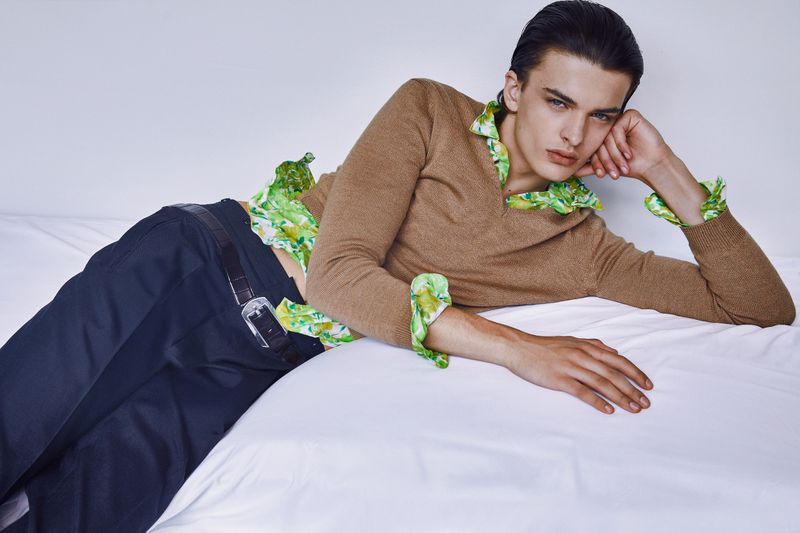 Male fashion model lying on a bed wearing a brown sweater and green floral shirt in a studio with soft, diffused lighting.