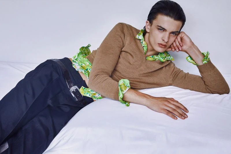 Male fashion model lying on a bed wearing a brown sweater and green floral shirt in a studio with soft, diffused lighting.