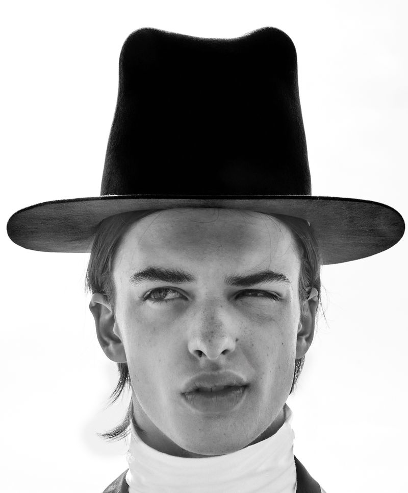 Monochrome portrait of a young adult with fair skin, wearing a black hat and a white turtleneck sweater against a light background.