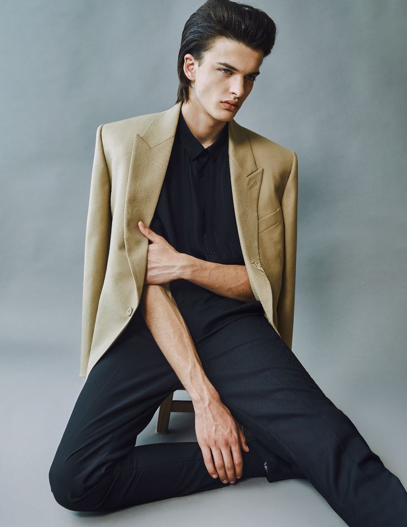 Portrait of a dark-haired male fashion model wearing a tan blazer and black shirt and pants, seated against a grey background in a photo studio.