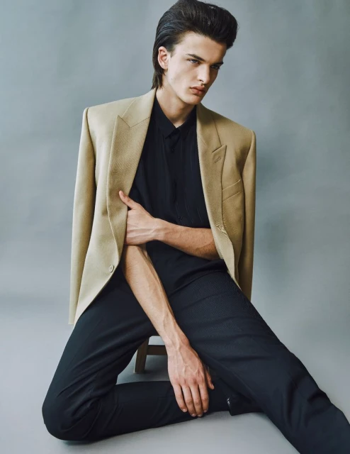 Portrait of a dark-haired male fashion model wearing a tan blazer and black shirt and pants, seated against a grey background in a photo studio.