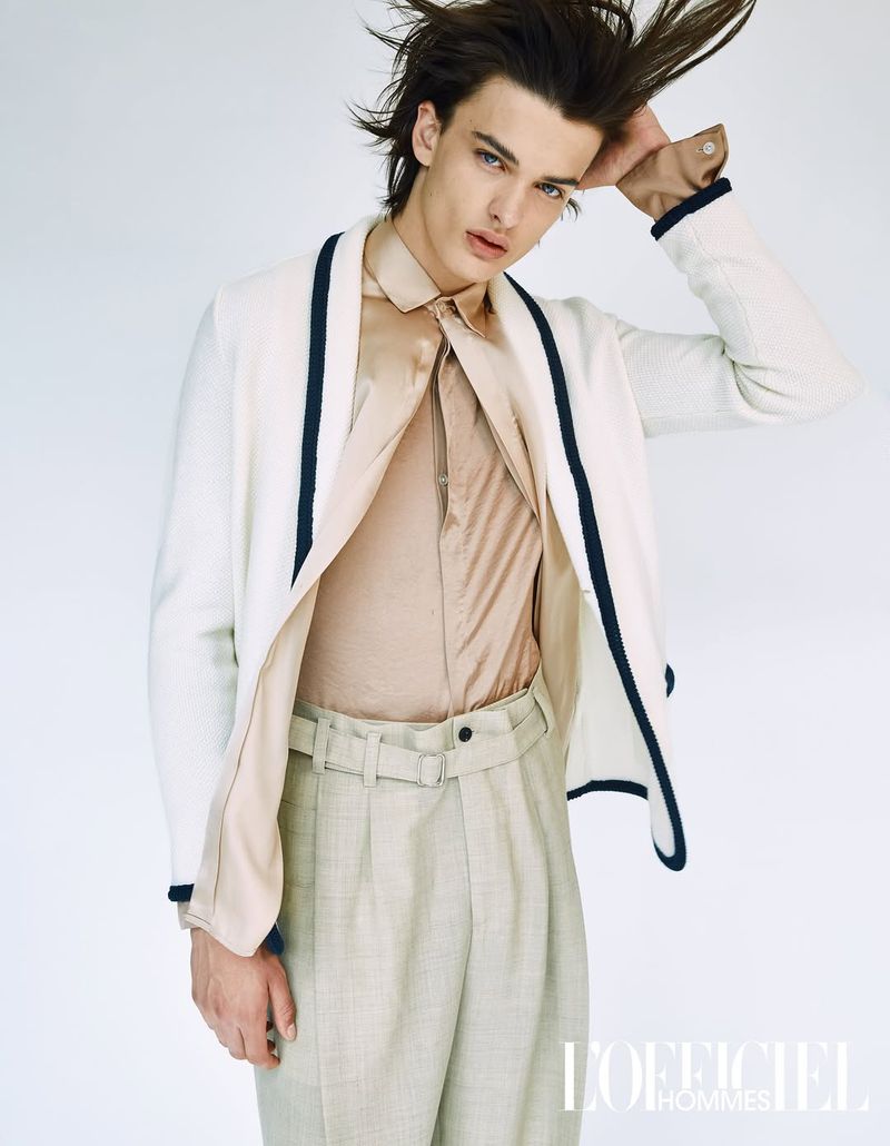 Portrait of a handsome male model in a silk shirt, cardigan and slacks posing in a photo studio, with the L'Officiel Hommes logo.