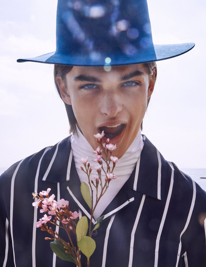 Portrait of a young man with a blue hat, striped shirt, and a turtleneck eating flowers, set against a bright, neutral background.