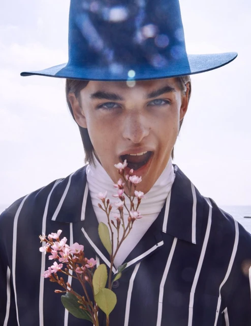 Portrait of a young man with a blue hat, striped shirt, and a turtleneck eating flowers, set against a bright, neutral background.