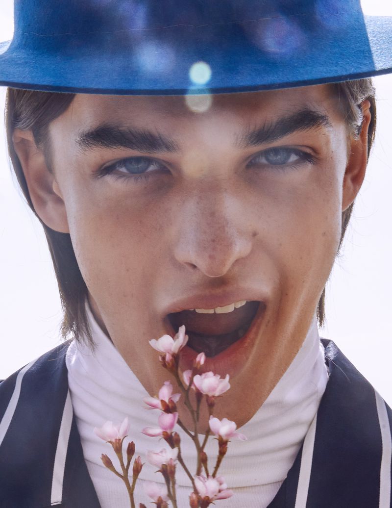 Close-up portrait of model Tristan Watkins wearing a blue hat and biting a small twig of pink blossoms for L'Officiel Hommes Ukraine.