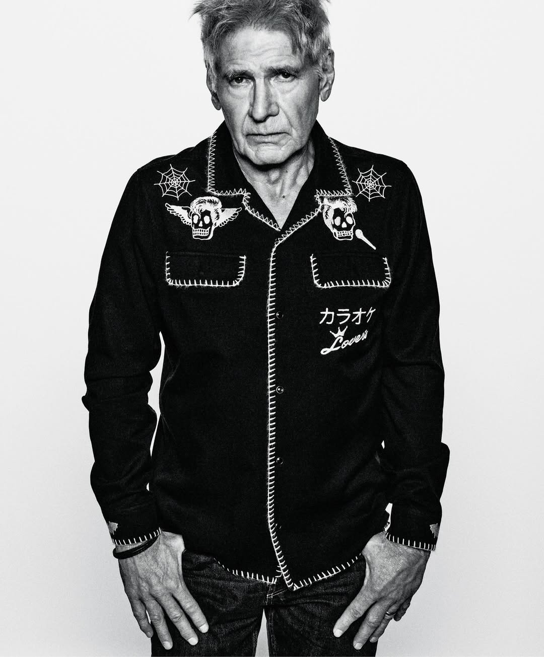 Monochrome portrait of Harrison Ford in a patterned black bowling shirt with skull and spiderweb graphics against a white background.