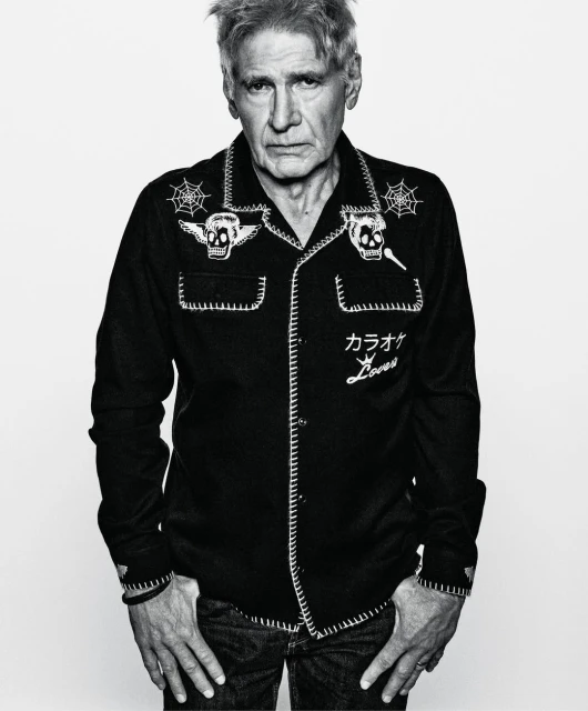 Monochrome portrait of Harrison Ford in a patterned black bowling shirt with skull and spiderweb graphics against a white background.