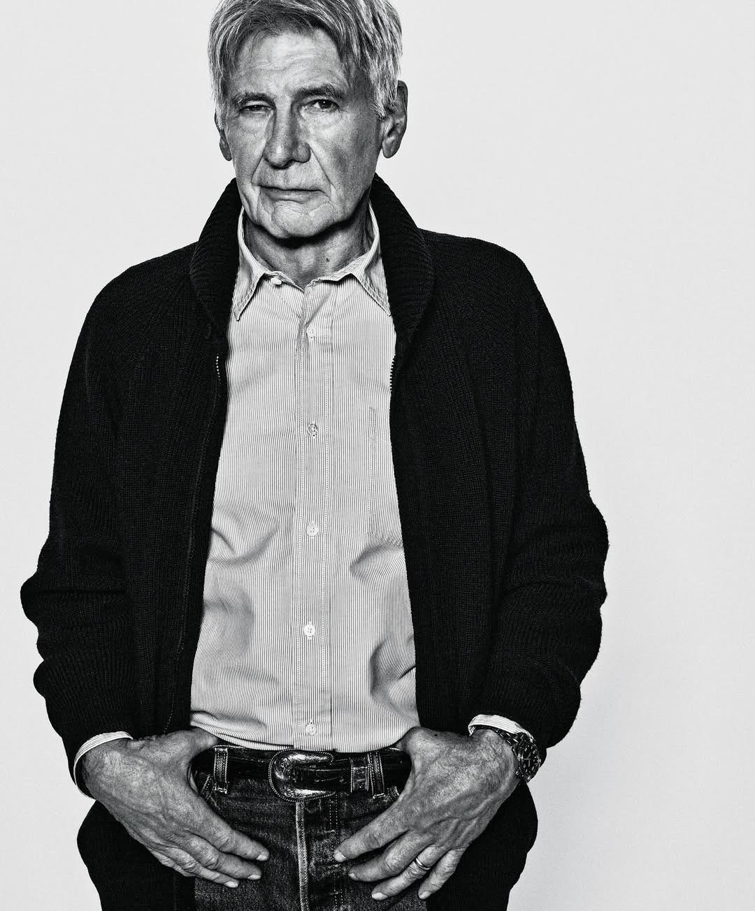 Black and white portrait of an older man in a shirt and cardigan, hands in his jean pockets, against a white backdrop.