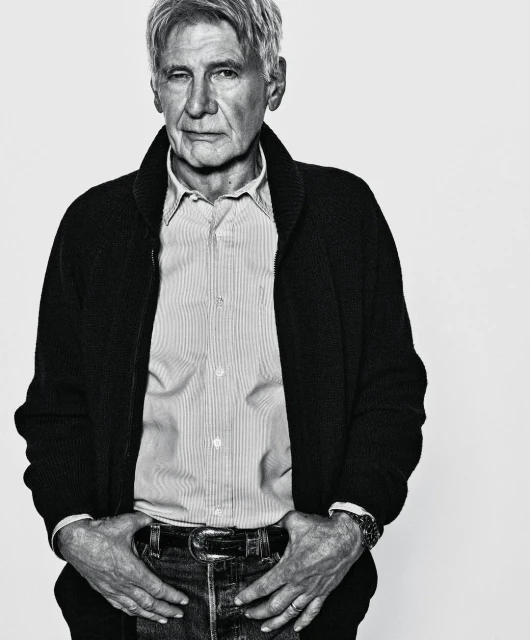 Black and white portrait of an older man in a shirt and cardigan, hands in his jean pockets, against a white backdrop.