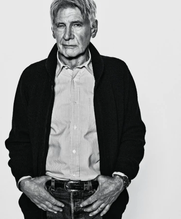 Black and white portrait of an older man in a shirt and cardigan, hands in his jean pockets, against a white backdrop.