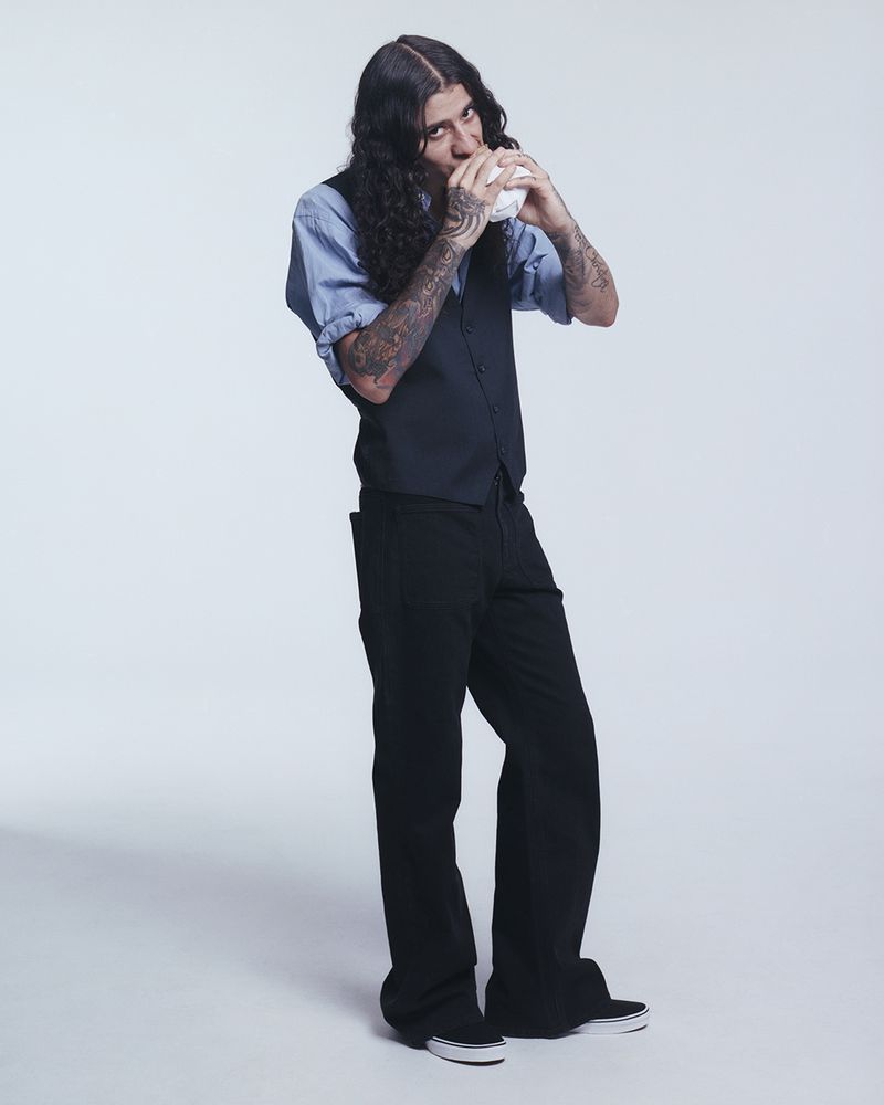 Man in a blue shirt, black vest, and black pants drinks from a white cup in a full-length studio shot against a white backdrop.