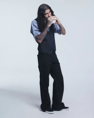 Man in a blue shirt, black vest, and black pants drinks from a white cup in a full-length studio shot against a white backdrop.
