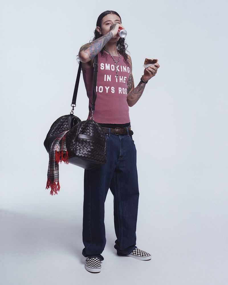 Man in a pink tank top drinks from a can while holding a pastry, wearing jeans, checkered Vans, and carrying a woven bag in a studio.