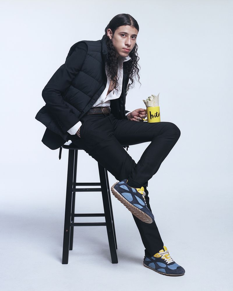 Man sitting on a stool in a photo studio, wearing a black puffer vest, white shirt, black pants, and blue and yellow athletic shoes.