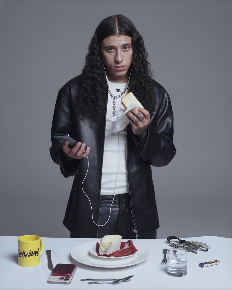 Young adult with long, wavy hair sits at a table with a phone, water glass, and cheesecake slice, wearing a black jacket and headphones.