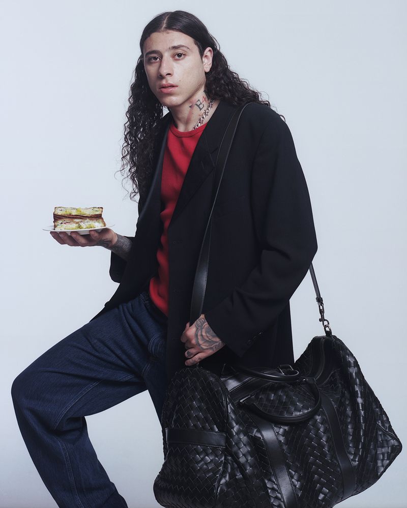 A young adult with long curly hair poses with a black blazer, jeans, a large woven leather bag, and holds a sandwich on a white background.