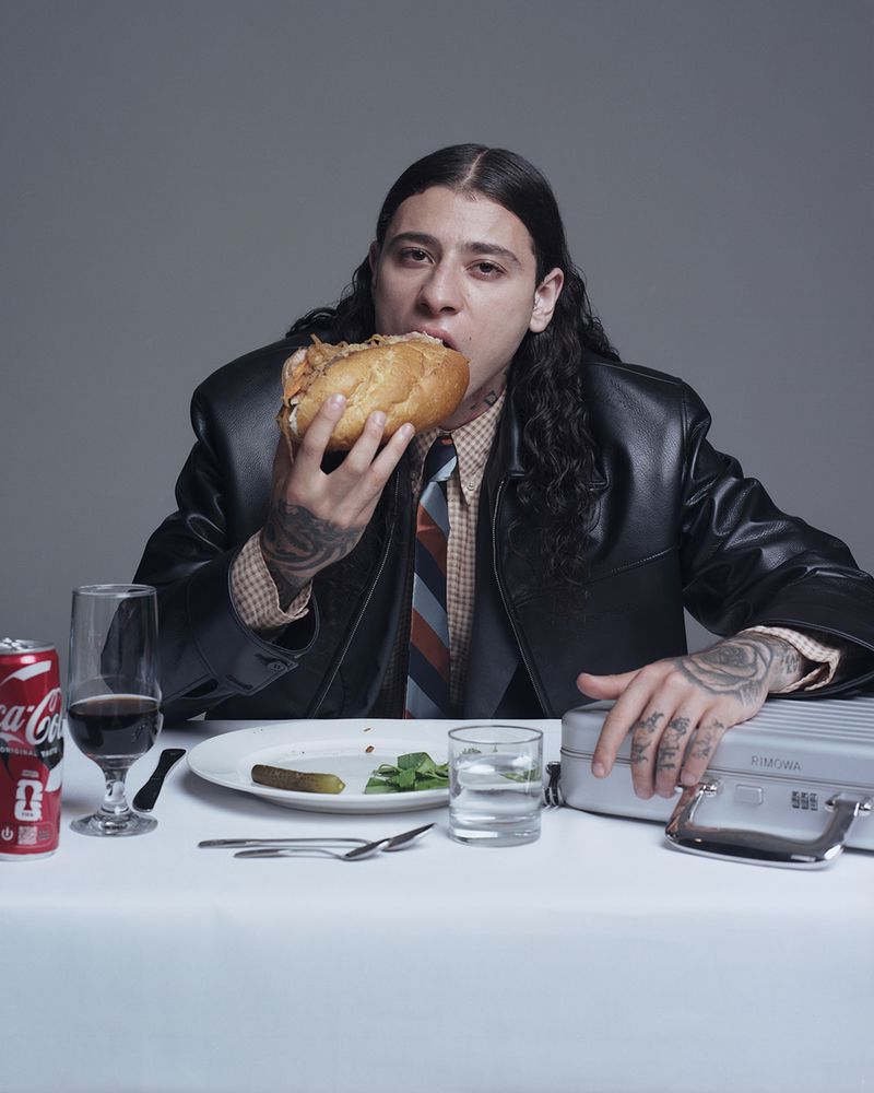 Adult man in a black jacket and patterned shirt eats a large sandwich at a table with a Coca-Cola can and silver briefcase.
