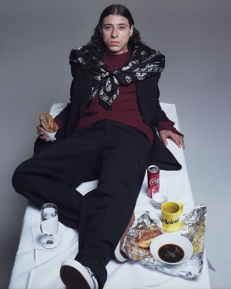 A young adult with long dark hair poses with food and drink on a white surface with grey backdrop, wearing a sweater, suit, and bandana.