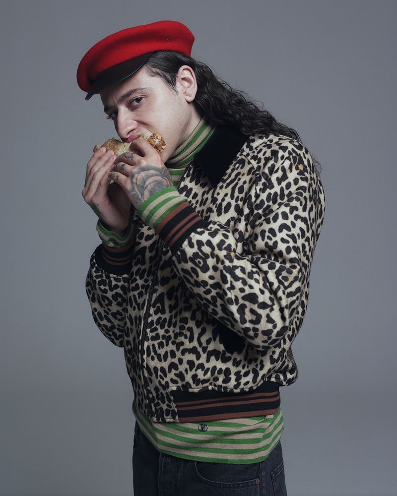 Man in a red beret and patterned jacket poses indoors while eating a pastry, showcasing fashion and lifestyle in a neutral setting.
