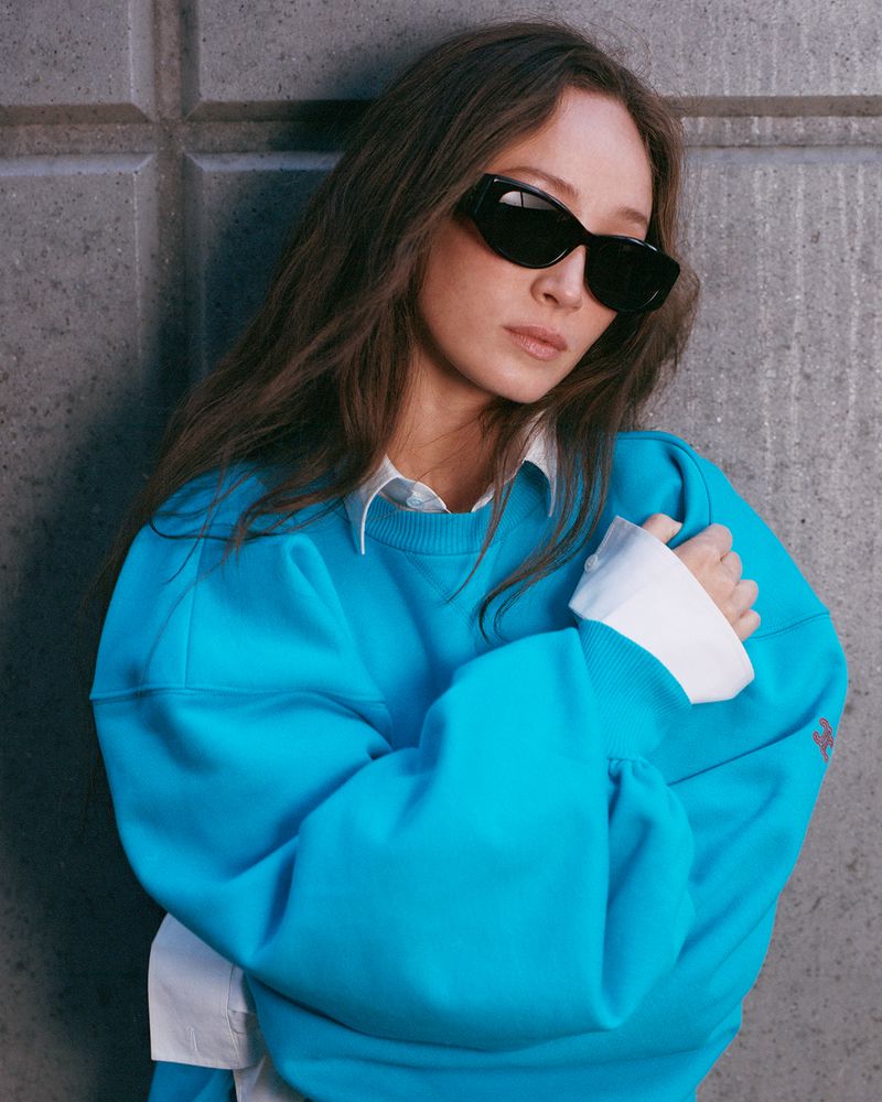Woman with long brown hair wearing sunglasses, a teal sweatshirt and white collared shirt, posing against a textured grey wall.