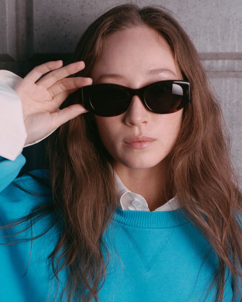 Portrait of a young woman wearing sunglasses, a blue sweatshirt and a white collared shirt, against a neutral wall.