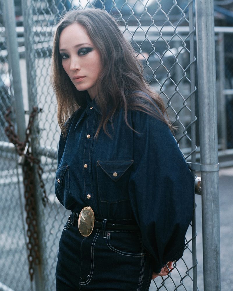 Stylish brunette woman in a denim outfit poses confidently in front of a chain-link fence, her gaze directed towards the viewer.
