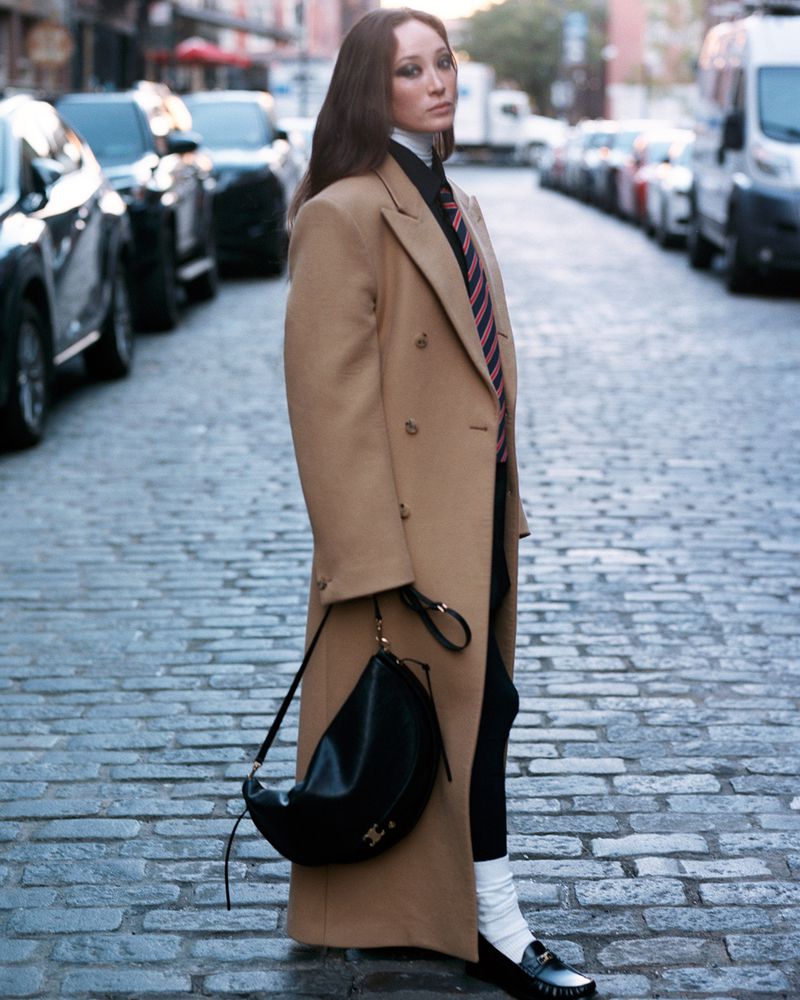 An adult female is seen in a full shot on a city street wearing a tan overcoat, black pants, and loafers with a black purse over her shoulder.