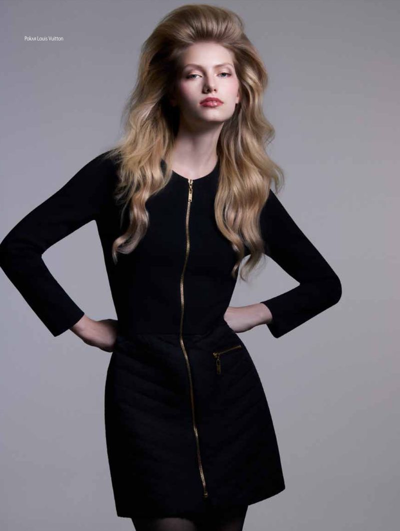 Fashion portrait of a fair-skinned adult female model with long blonde hair wearing a black zip-front dress against a gray background.