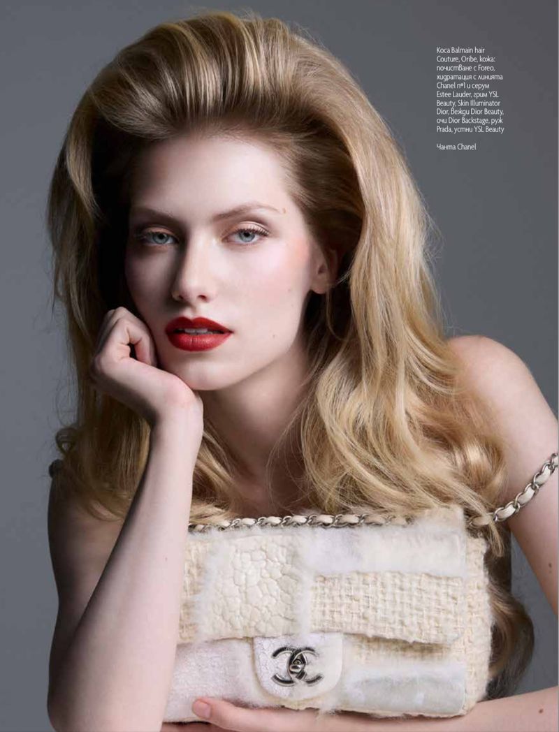 Blonde woman poses in photo studio with Chanel handbag. The subject is framed in a medium shot against grey backdrop, wearing red lipstick.