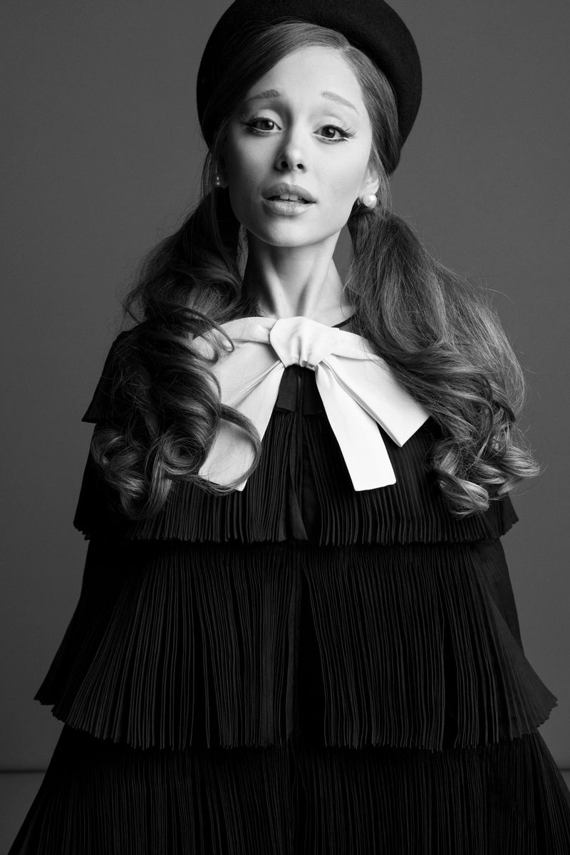 Black and white portrait of a woman with pigtails and a beret, wearing a tiered dress with a large bow, against a grey background.