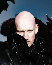 Bald man wearing a black leather jacket with an exaggerated upturned collar and fur trim against a muted blue backdrop.