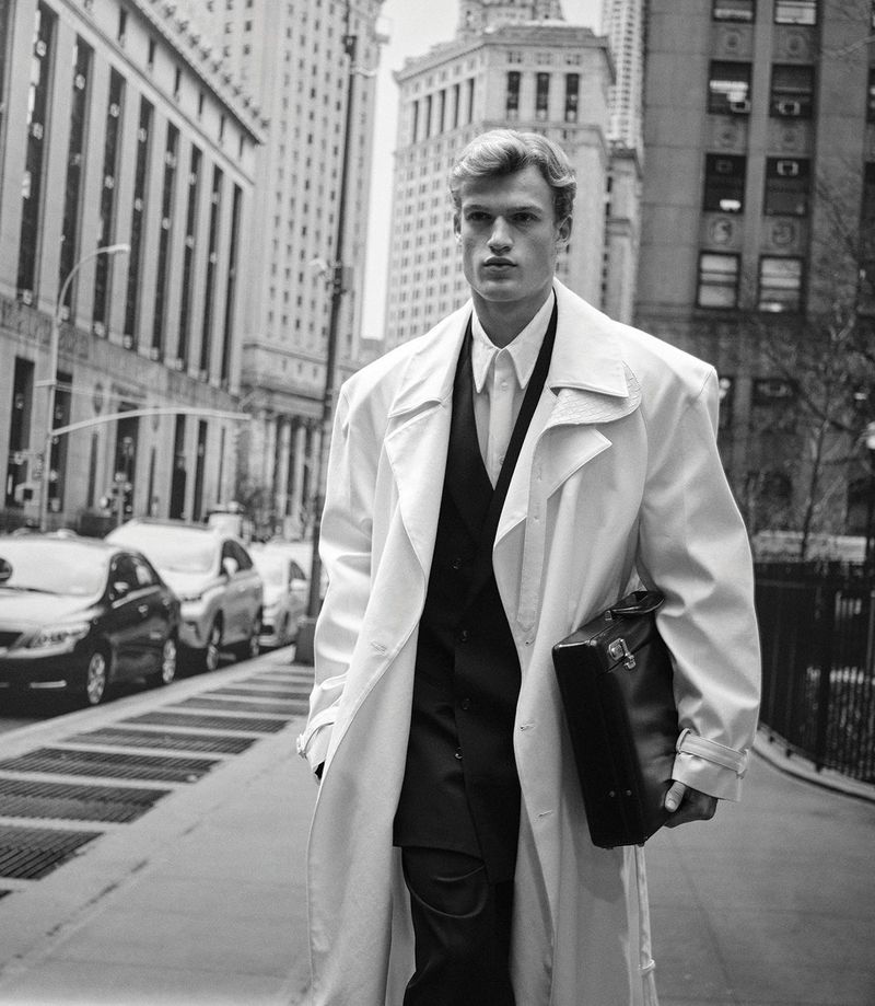 A blond male model in a white trench coat walks on a city sidewalk in black and white, carrying a briefcase, tall buildings in the background.
