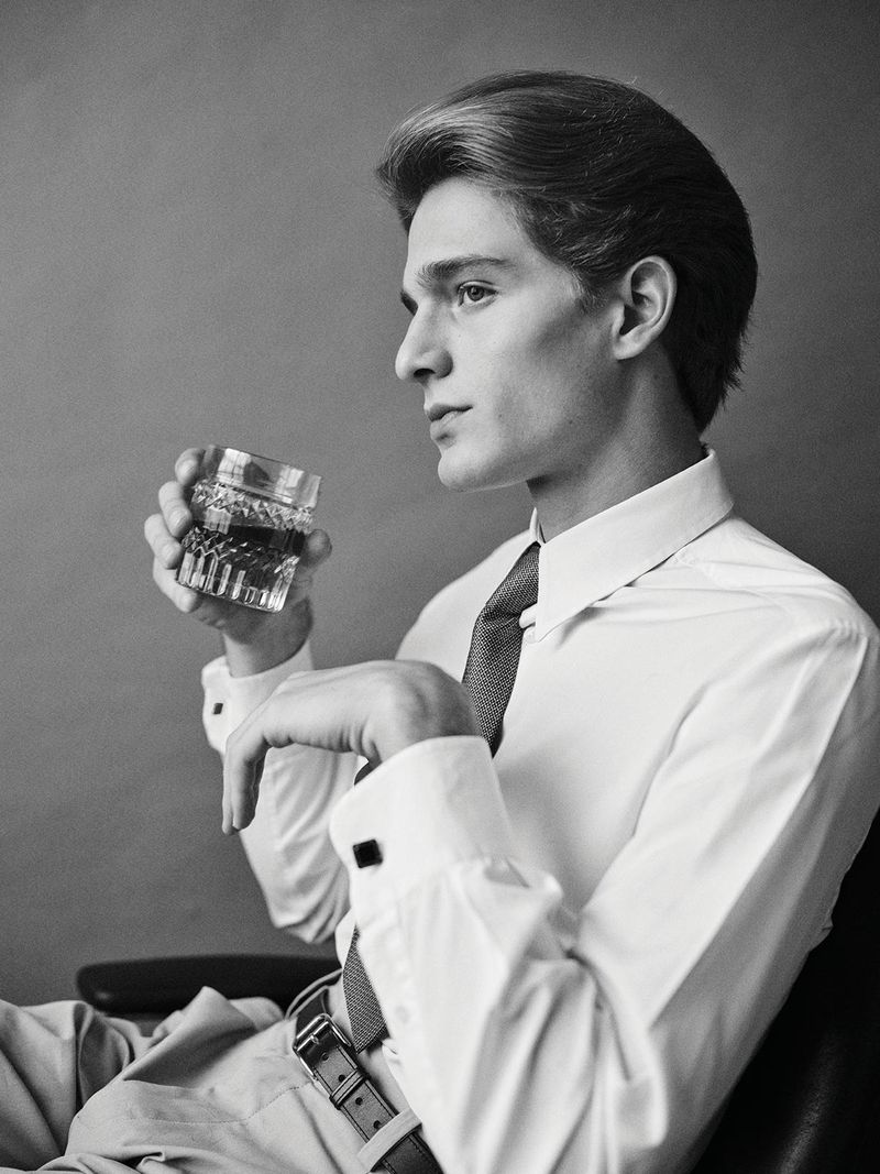 Black and white portrait of a young adult male in a shirt and tie, holding a faceted glass of liquor while seated indoors.