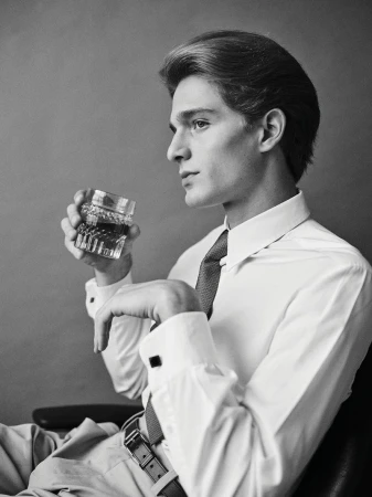 Black and white portrait of a young adult male in a shirt and tie, holding a faceted glass of liquor while seated indoors.