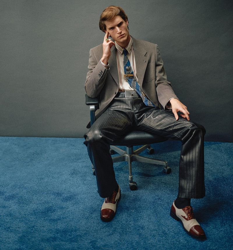 Adult male sits in office chair wearing a grey suit, patterned shirt, and leather pants in an indoor studio environment with blue flooring.