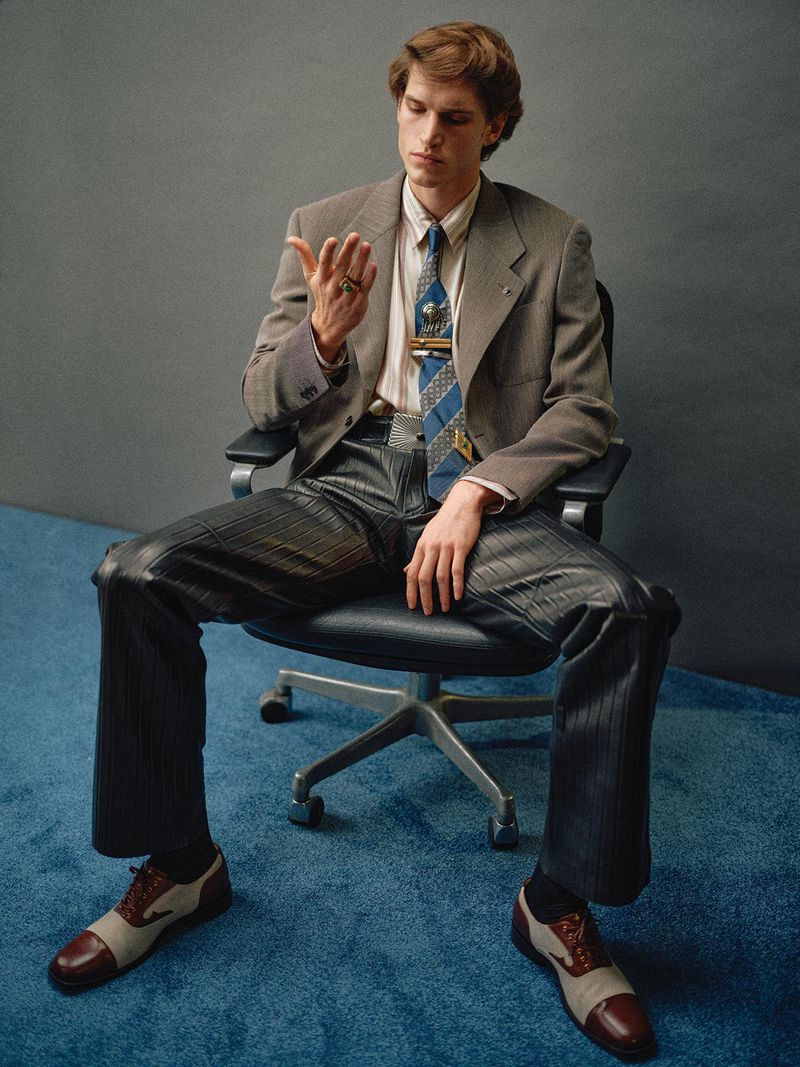 Portrait of a young man seated in an office chair, wearing a suit jacket, tie, and leather pants. Professional fashion editorial.
