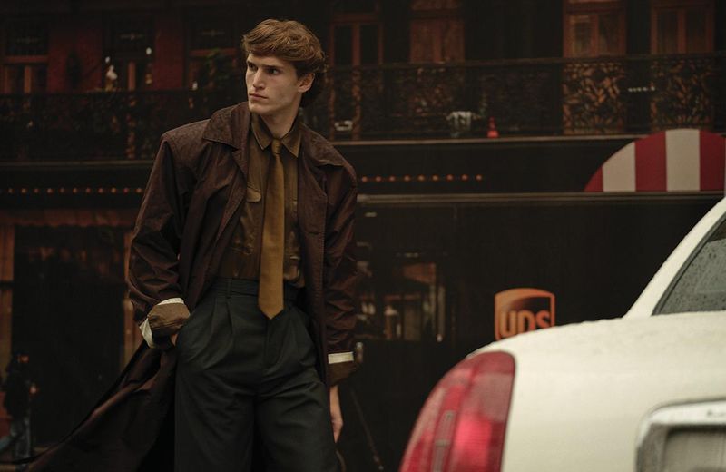 Male model poses in a brown trench coat and dark trousers next to a white car against a dark urban backdrop, stylish and fashionable.