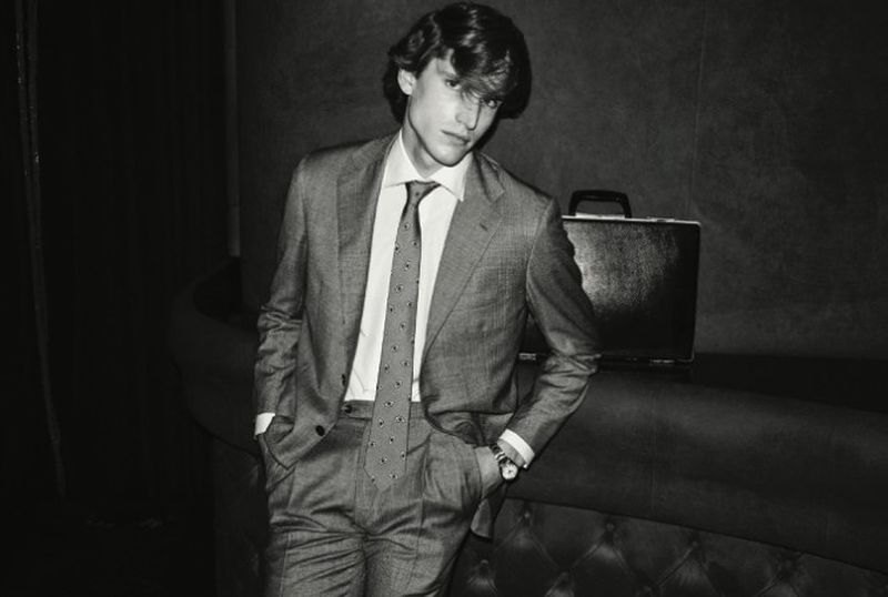 Monochrome portrait of an adult male in a suit with a briefcase, leaning against a sofa in an indoor setting with dark lighting.