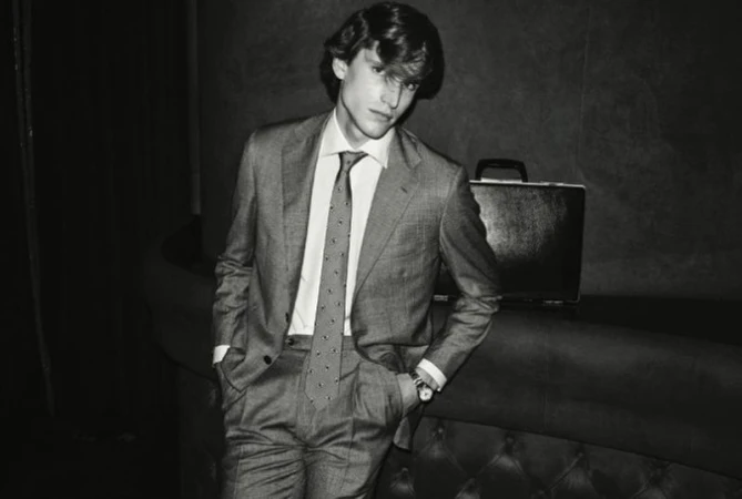 Monochrome portrait of an adult male in a suit with a briefcase, leaning against a sofa in an indoor setting with dark lighting.