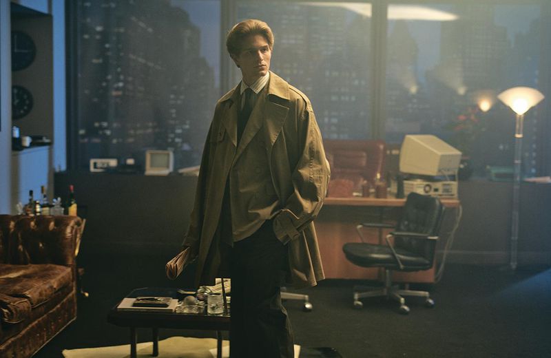 Young man in a trench coat stands in a vintage-style office with a city view, dimly lit by desk lamps and overhead fixtures.