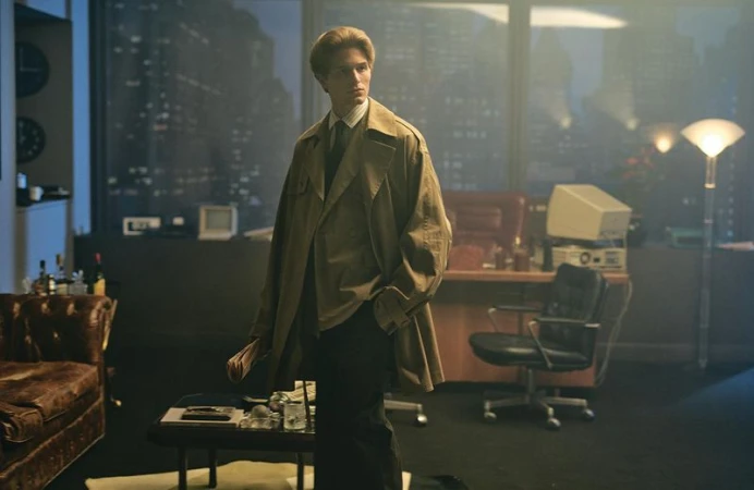 Young man in a trench coat stands in a vintage-style office with a city view, dimly lit by desk lamps and overhead fixtures.