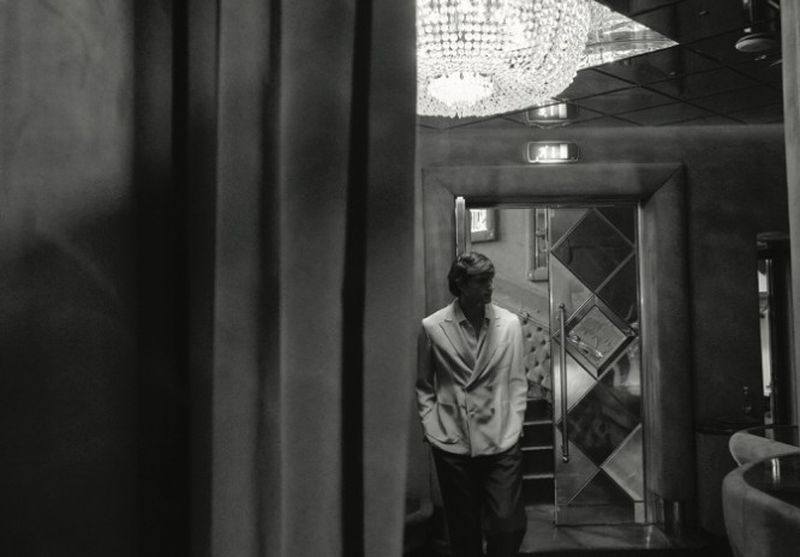 Black and white film photo of an adult male in a suit, indoors, walking toward a door in a textured, vintage-style entertainment venue.