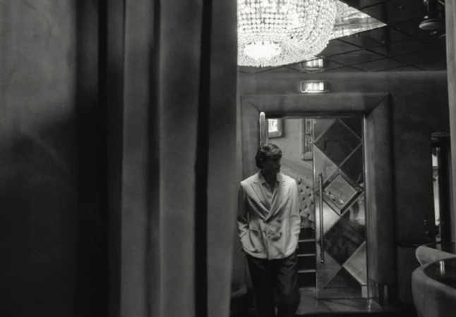 Black and white film photo of an adult male in a suit, indoors, walking toward a door in a textured, vintage-style entertainment venue.