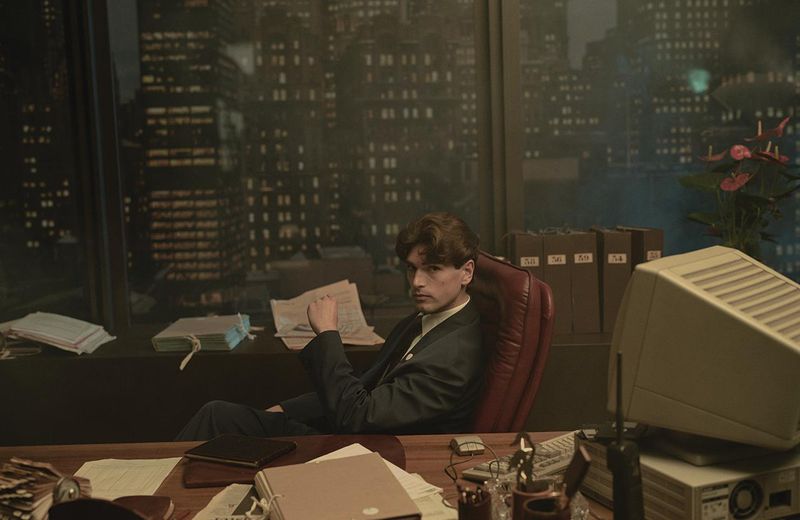 Man in a suit sits at a cluttered office desk in front of a city skyline at night, illuminated by dim, vintage-style lighting.
