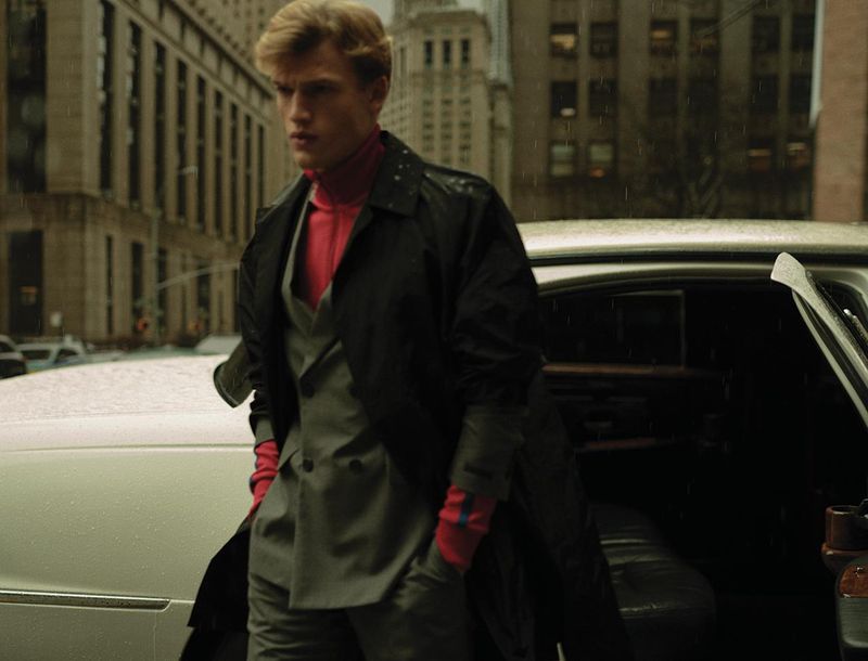 Fashion shot of a man in a suit and trench coat exiting a limousine in a city environment on an overcast day with nearby buildings.