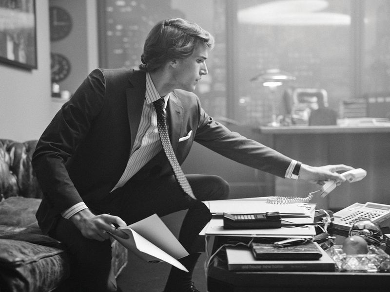 Black and white shot of a businessman in a suit sitting on a leather couch reaching for a telephone receiver in an office setting.